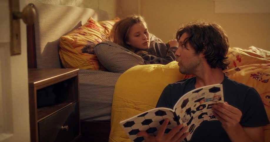 A loving father reads a soccer-themed book to his young daughter as she rests in her sunlit bed