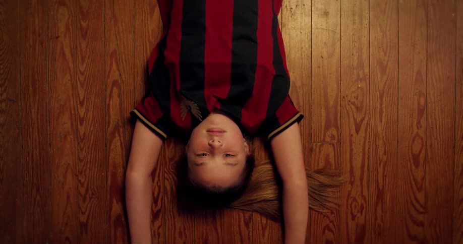 From a top-down perspective, a person pulls a young girl by her arms across a wooden floor until she comes into full view