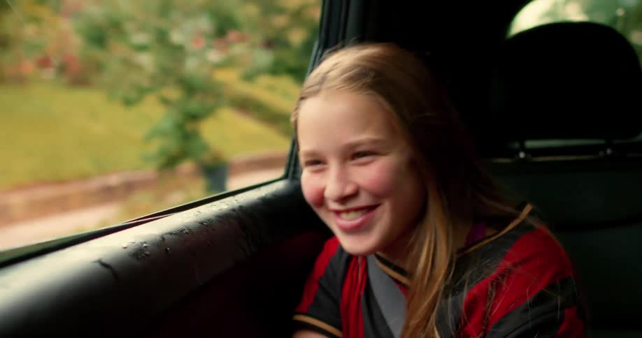 A happy young girl in a soccer jersey laughs and smiles while riding in the back of a car in slow motion