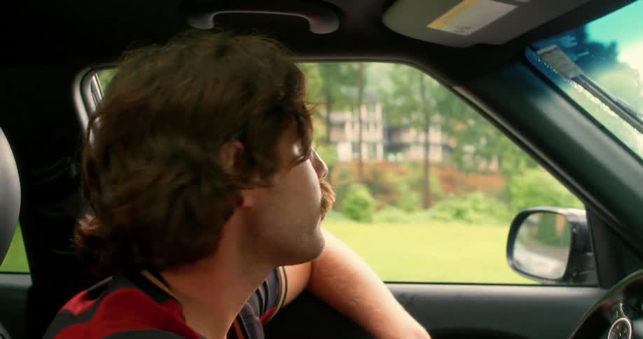 Close-up of a man with a stylish mustache and mullet driving his car through a residential neighborhood in slow motion