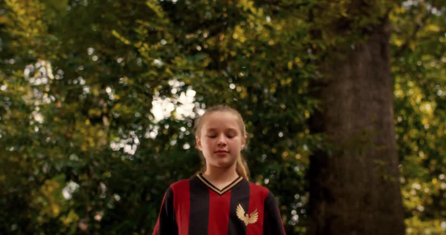 A low angle slow motion shot of a young girl in a soccer jersey taking a deep breath and focusing in a park