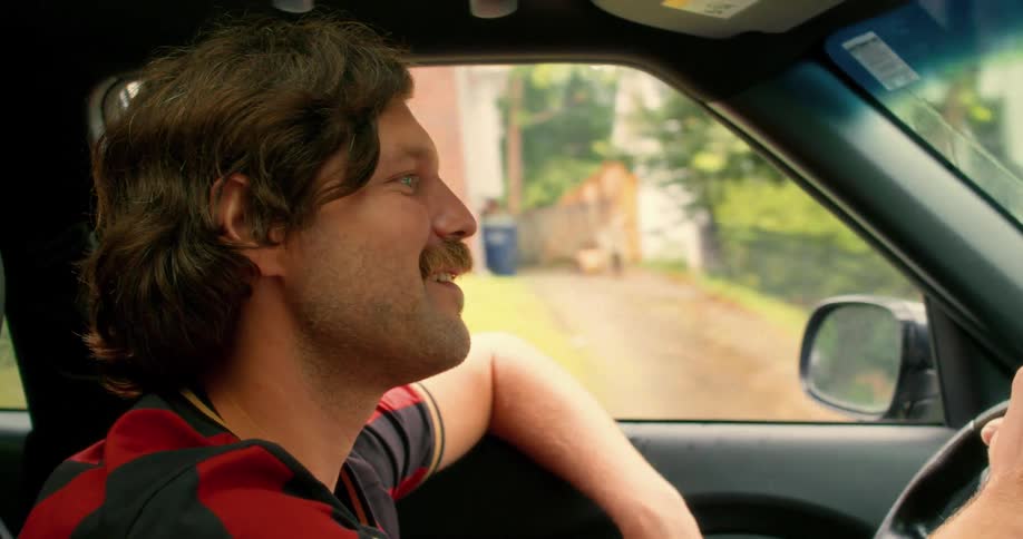 A cheerful man with a retro hairstyle and mustache happily drives his car through a suburban neighborhood in slow motion