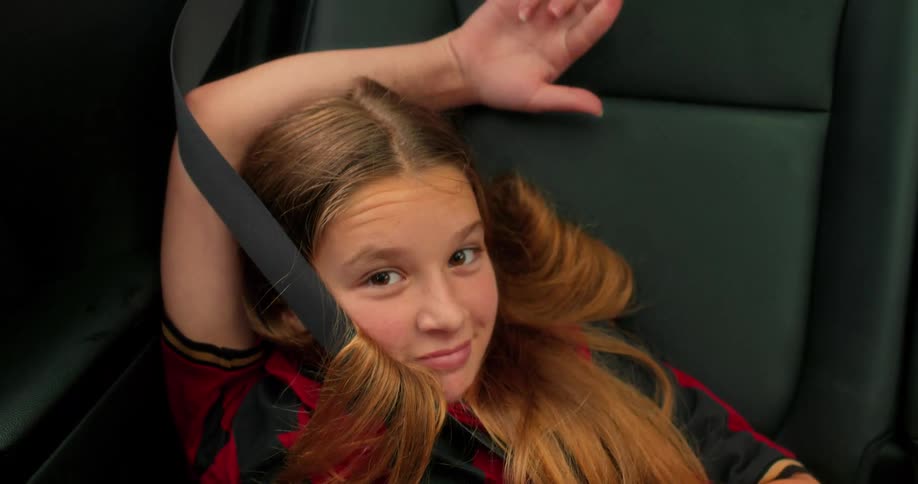 A happy young girl with long hair smiles and laughs while buckled into the backseat of a car