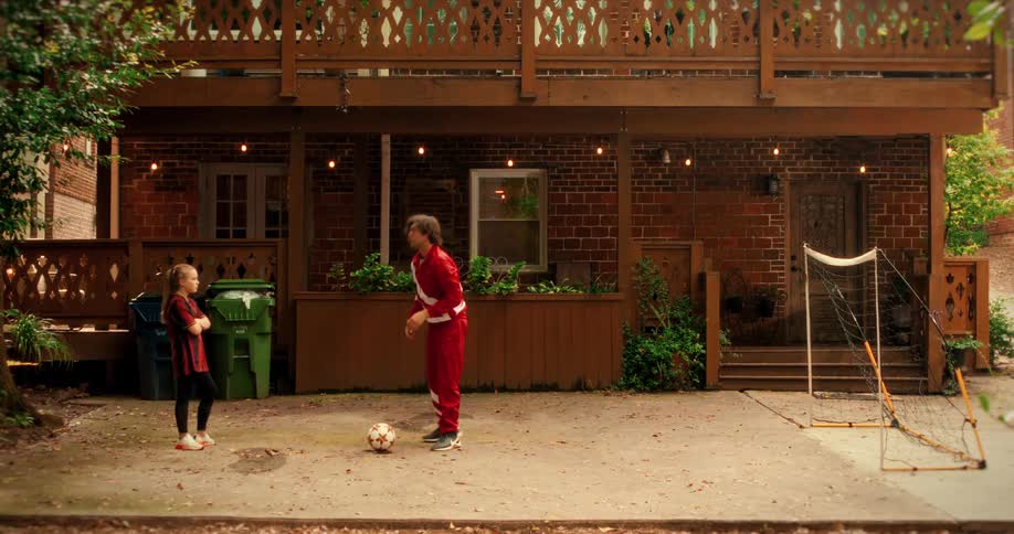 A father in a red and white tracksuit coaches his young daughter in soccer in their backyard driveway