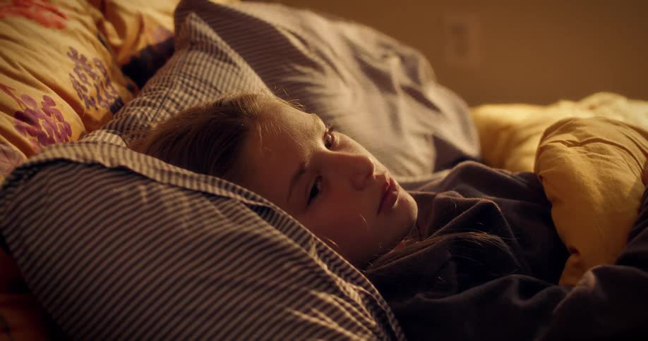 A young girl bathed in warm morning light sleeps peacefully in her cozy bed before slowly waking up