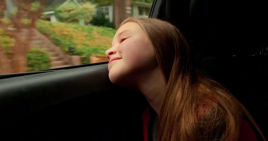 A serene young girl with long hair smiles and rests her head while looking out the window of a moving car
