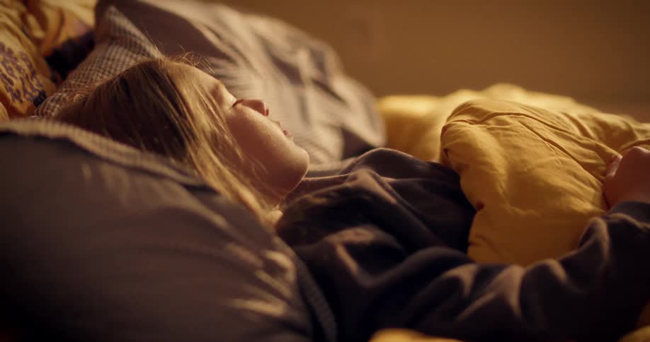 A young woman sleeps peacefully in her cozy, warm-lit bed, tucked under a yellow comforter