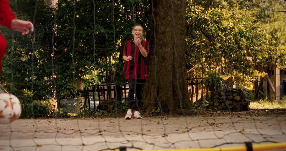 A young girl cheers and jumps for joy in slow motion as her father scores a goal while they practice soccer together outdoors