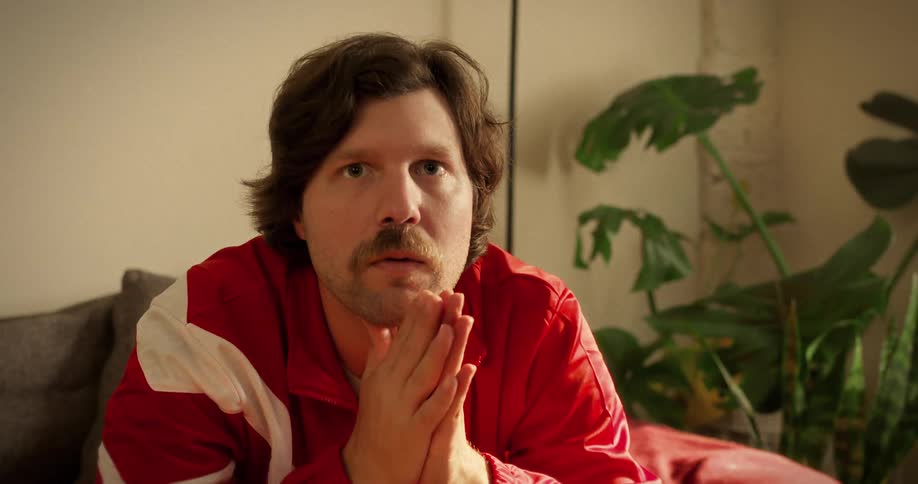 A man with a mustache in a red tracksuit has an intense emotional reaction while watching a tense game at home