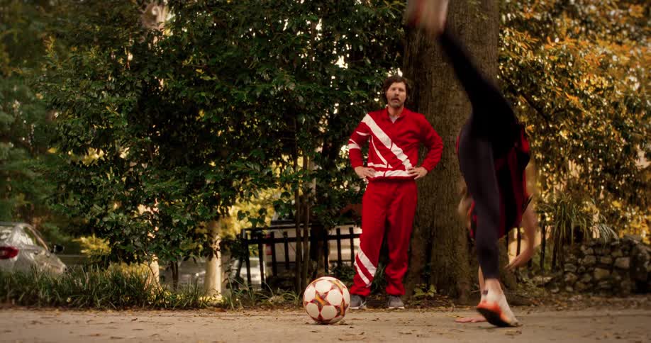 A talented young girl in a red and black soccer jersey performs impressive cartwheels and gymnastic tricks in a park while her coach watches in slow motion