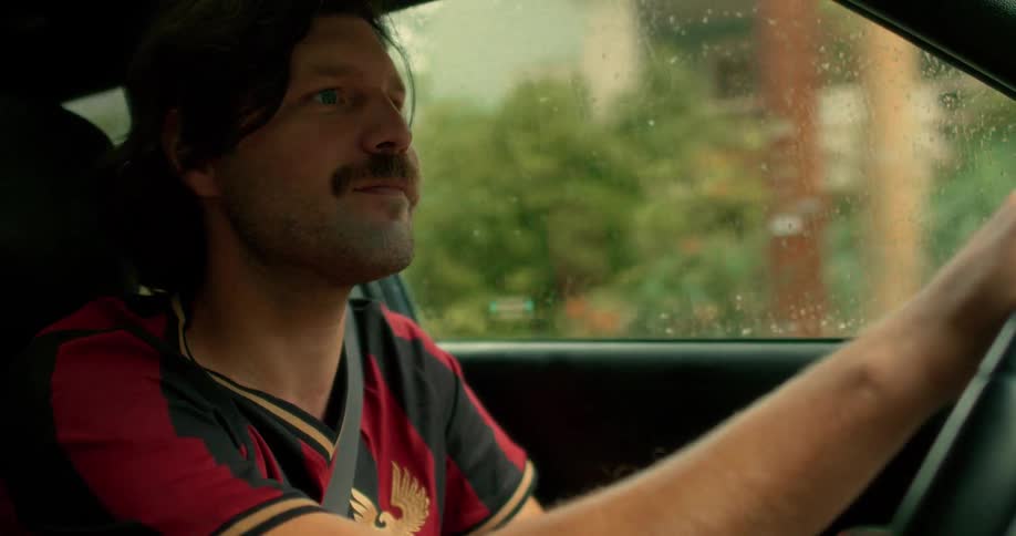 A focused man with a vintage mustache drives his car through a neighborhood on a rainy day