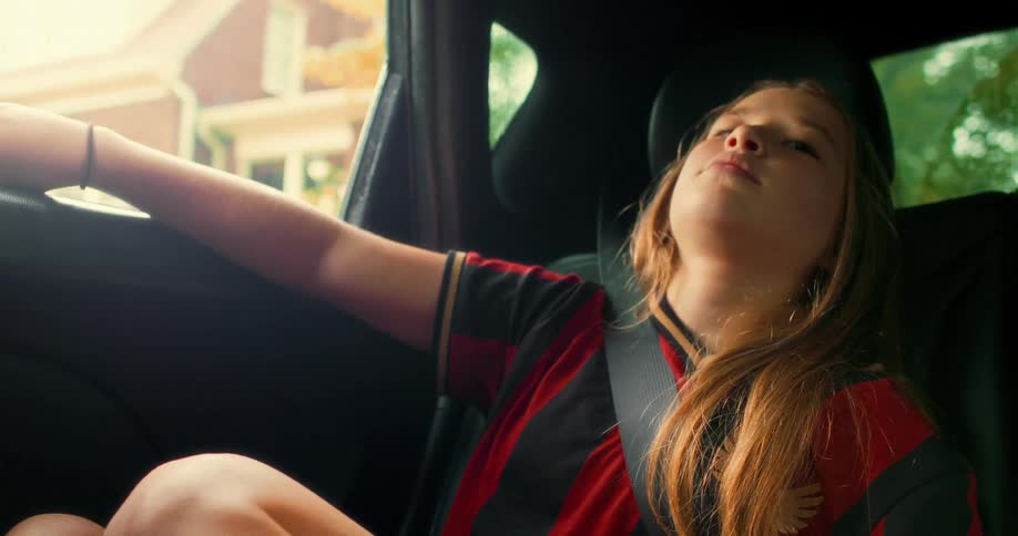 A relaxed young girl in a soccer jersey rests in the back of a moving car, enjoying the ride