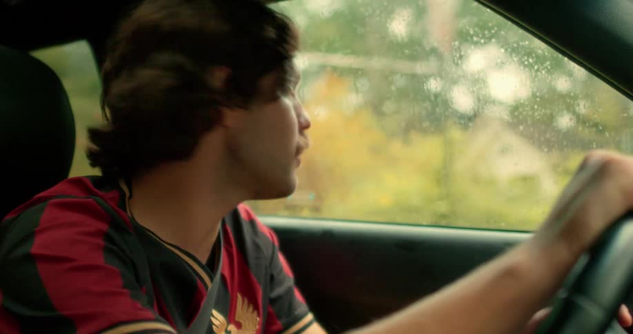A man with a mustache wearing a soccer jersey talks while driving his car on a rainy day