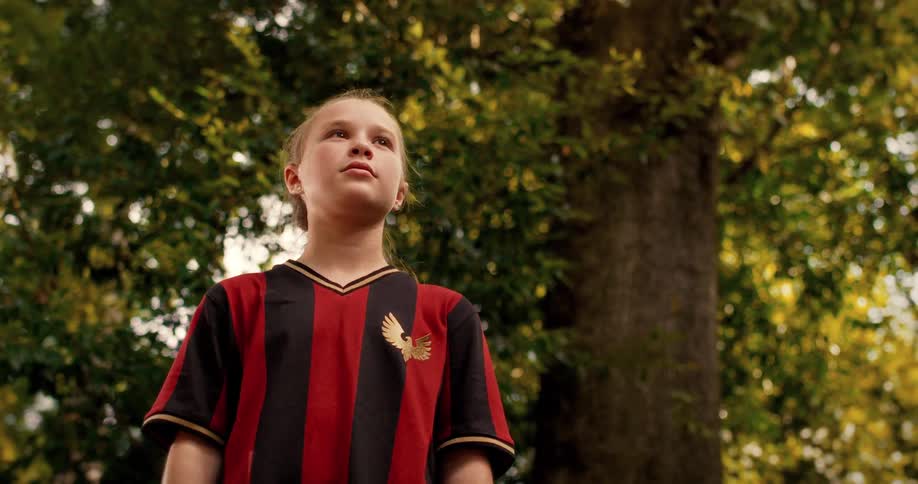 An inspired young girl in a soccer jersey stands with focus and determination under a large tree