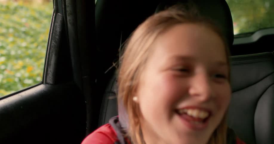 Slow motion close-up of a happy young girl laughing hysterically and covering her face while riding in the back seat of a car