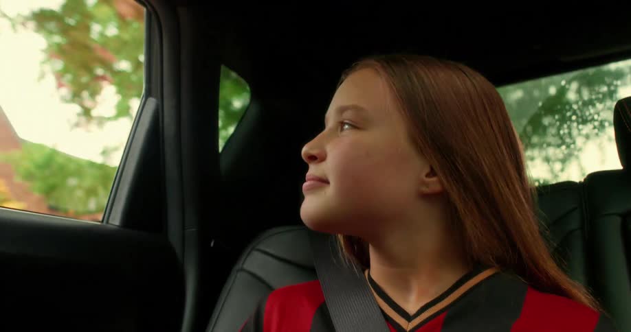 Content young girl in a soccer jersey smiles while gazing out the window during a car ride in slow motion