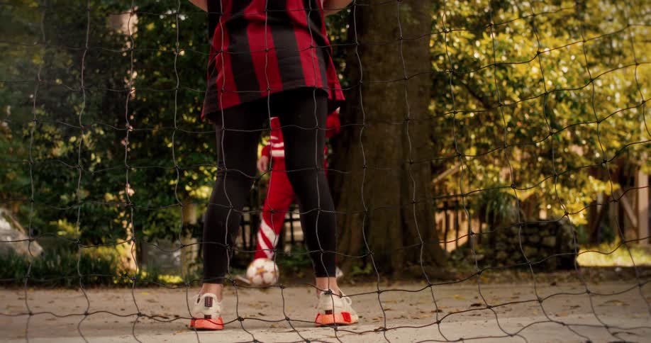 A man in a red tracksuit practices his soccer moves against a woman goalie in slow motion, shot from behind the net