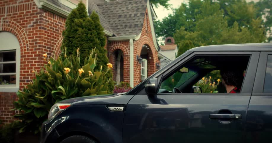 A man in a soccer jersey slowly pulls up in his car and gets out in front of a beautiful brick suburban home
