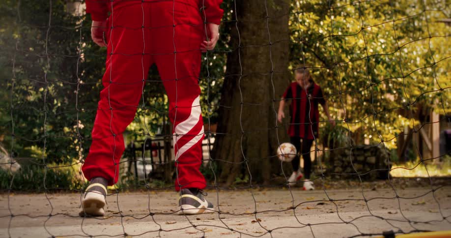 From behind the net, a young girl skillfully dribbles a soccer ball towards a boy playing goalie in their backyard in slow motion
