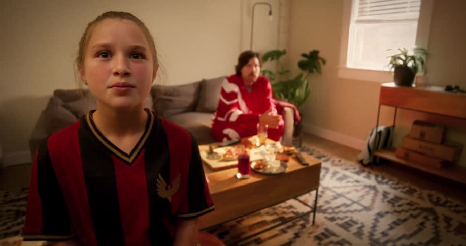 A young girl in a soccer jersey watches a tense game on TV while her father reacts with disappointment in the background