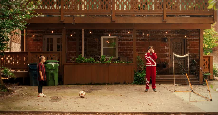 A father in a silly red tracksuit coaches his young daughter during soccer practice in their backyard, and she scores a goal