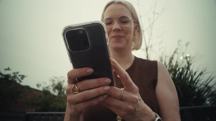 A slow push-in, low-angle shot of a smiling woman using her smartphone on a park bench on a cloudy day