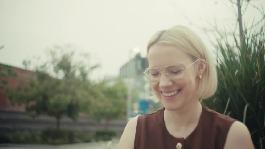 A stylish blonde woman with glasses smiles while using her smartphone on a bench in a modern city park
