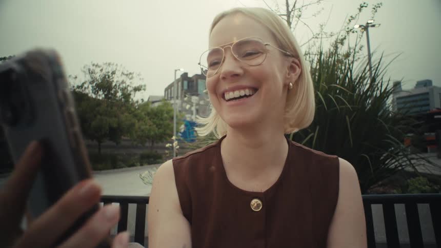 A smiling woman with glasses enjoys a cheerful video call on her smartphone, waving happily while sitting outside in the city