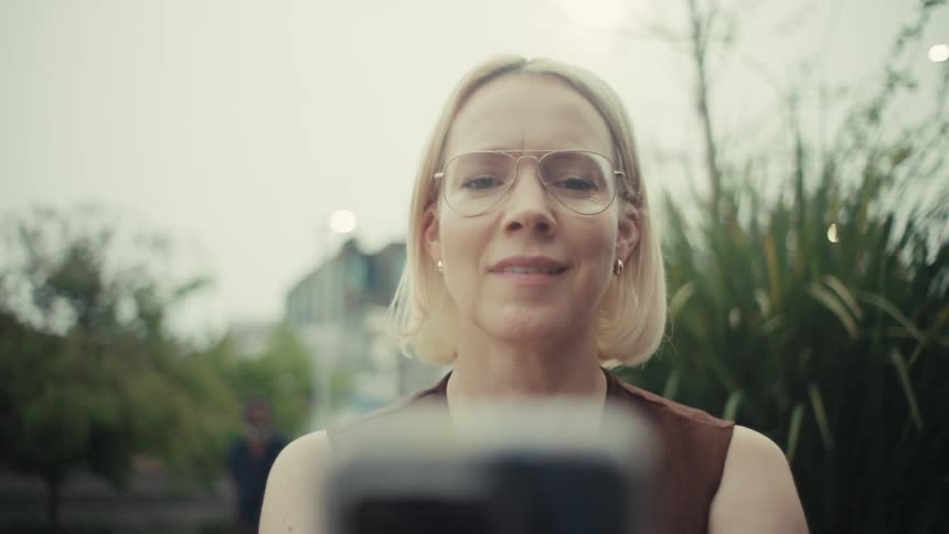 A stylish blonde woman wearing glasses smiles as she looks at her phone while sitting outside in the city