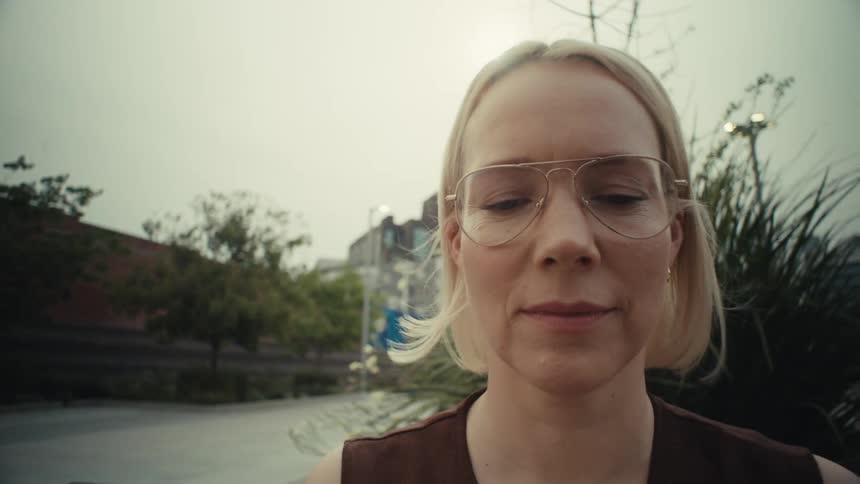 Slow motion pull-out shot of a pensive blonde woman with glasses sitting on a park bench and looking at her phone