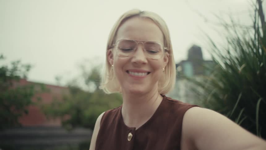 A stylish blonde woman with glasses sitting on a park bench smiles after checking her smartphone and smartwatch