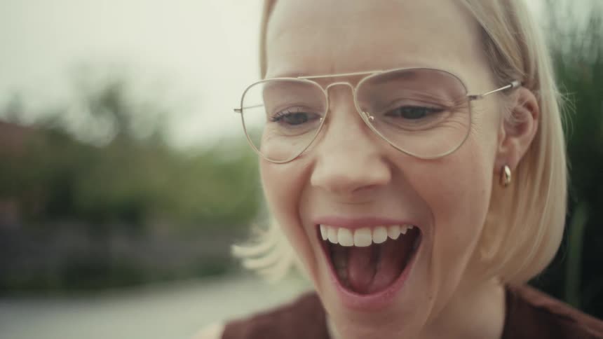 A stylish blonde woman reacts with joyful surprise and an open mouth while looking at her phone in a slow-motion close-up