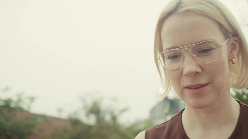 A smiling blonde woman with glasses texts and laughs while using her smartphone on an outdoor bench in slow motion