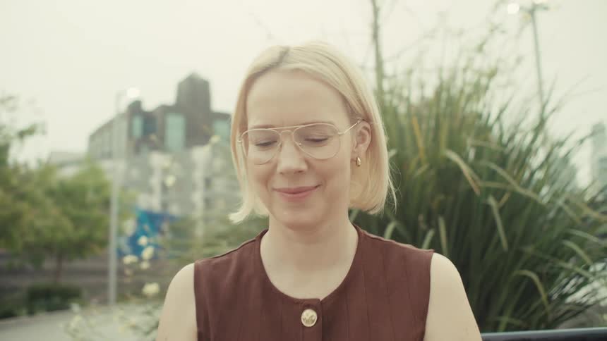 A stylish blonde woman with glasses smiles as she uses her smartphone while sitting on a bench in a city park