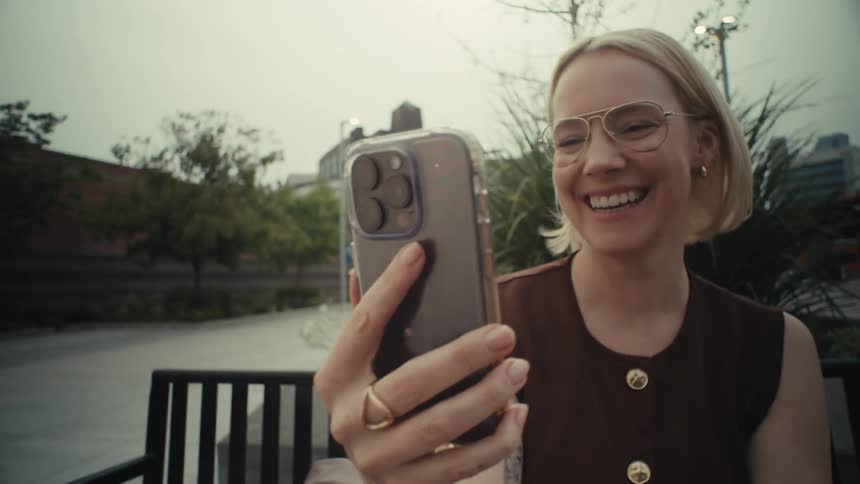 A joyful blonde woman with glasses smiles and waves goodbye while on a video call on her smartphone in an urban park