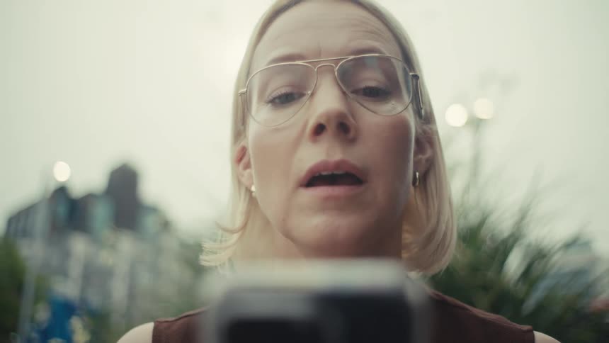 An emotional close-up shows a woman's face change from worry to relief and happiness as she reads something on her phone in an urban park