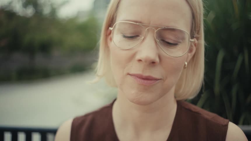 A thoughtful blonde woman in glasses sits on a park bench and uses her smartphone in slow motion
