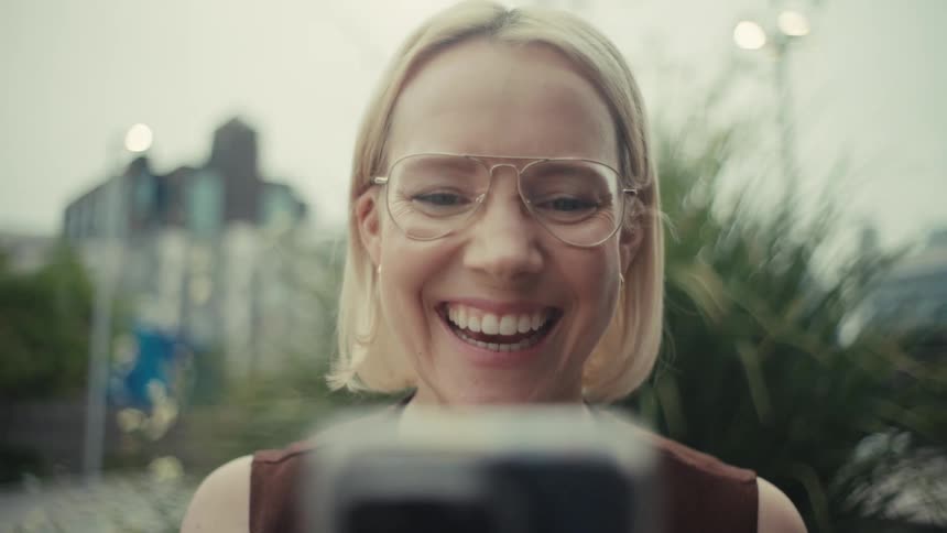 A stylish blonde woman in glasses laughs and waves enthusiastically during a video call on her phone outdoors