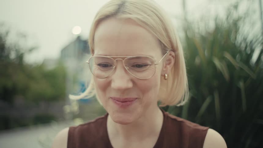 A joyful blonde woman in glasses watches her phone and bursts into laughter in a slow-motion close-up