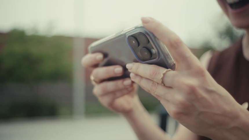 A joyful blonde woman wearing glasses laughs heartily as she watches an entertaining video on her smartphone outdoors in slow motion
