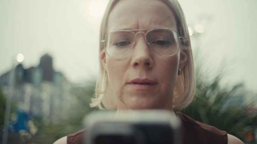 A close-up of a blonde woman in glasses looking at her phone, her serious expression breaking into a joyful smile