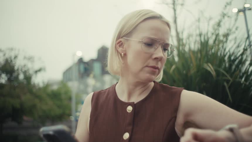 A professionally dressed woman checks her smartphone and smartwatch while waiting on a bench in a modern urban park