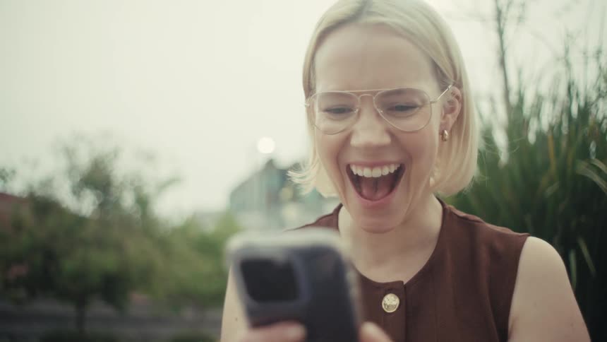 A stylish blonde woman sitting on a city bench erupts in pure joy and excitement while looking at her smartphone