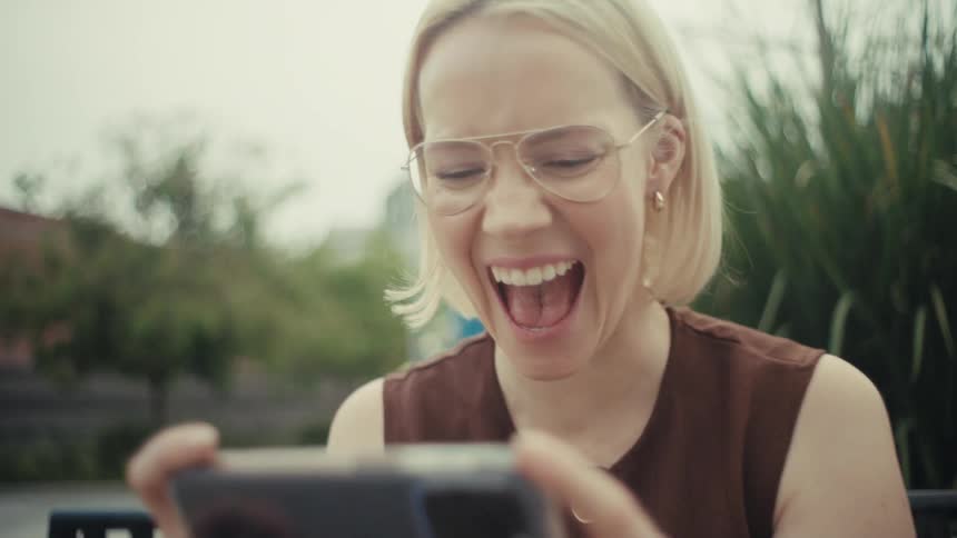 A stylish blonde woman sitting on a park bench laughs uncontrollably while watching something on her phone in slow motion