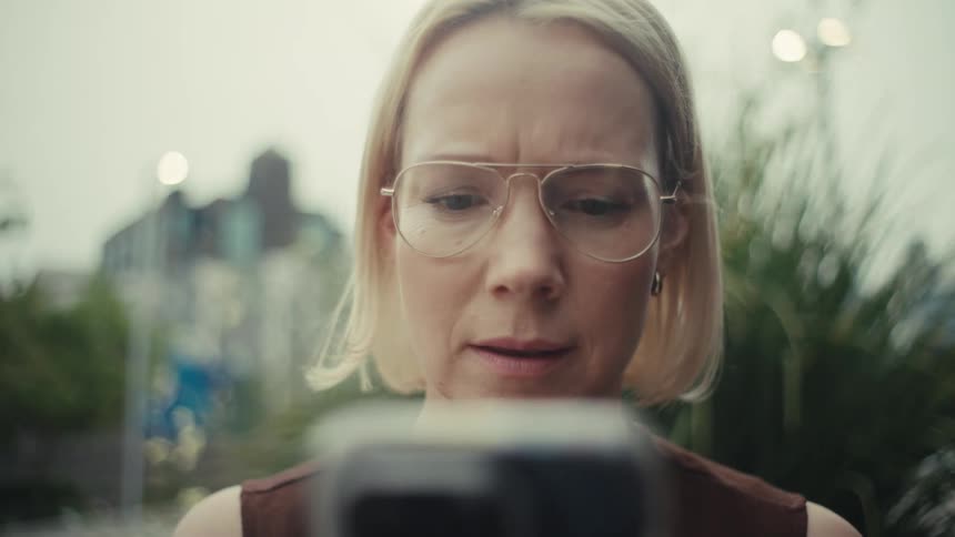 A slow-motion close-up shot of a serious blonde woman in glasses looking at her smartphone screen in a city park