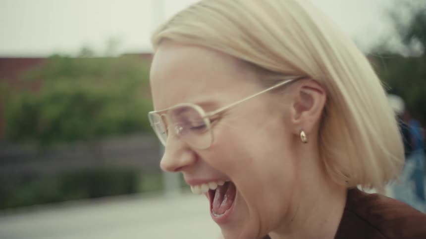 A stylish blonde woman with glasses laughs out loud in slow motion while watching something on her smartphone on a city bench