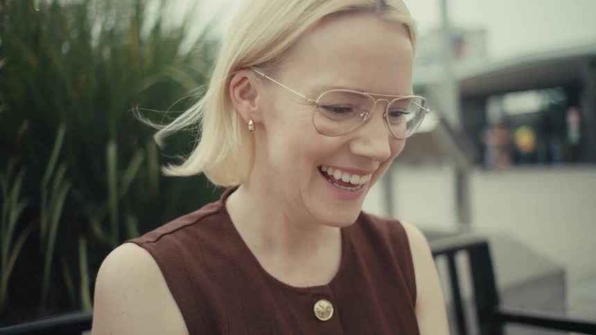 Slow motion shot of a happy woman with glasses laughing while watching a video on her smartphone on a city bench
