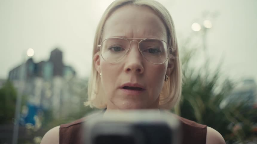 A close-up of a blonde woman wearing glasses, staring intently at her phone with a concerned and shocked expression while outdoors in the city