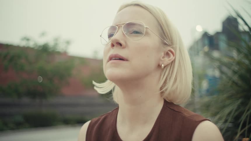 A thoughtful woman with glasses puts down her phone to take a reflective deep breath while sitting on a bench outdoors