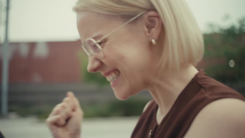 Slow motion shot of an ecstatic blonde woman celebrating winning a mobile game while sitting on a park bench